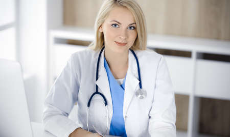 Portrate of woman-doctor at work while sitting at the desk in clinic. Blonde cheerful physician ready to help patientsの写真素材