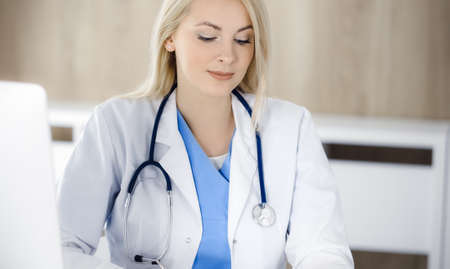 Woman-doctor at work while sitting at the desk in hospital or clinic. Blonde cheerful physician filling up medication history record formの写真素材