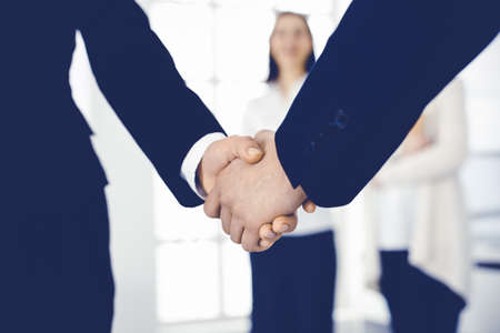 Businessman and woman shaking hands with colleagues at the background. Handshake at meeting in...