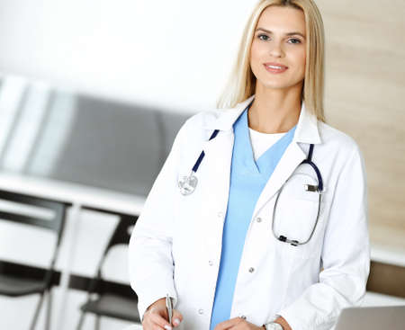 Doctor woman at work in hospital excited and happy of her profession. Blonde physician controls medication history records and exam results while using clipboard. Medicine and healthcare conceptの写真素材