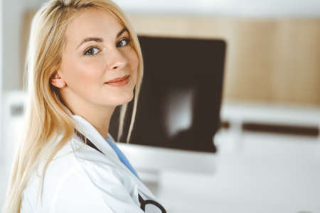 Portrate of woman-doctor at work while sitting at the desk in clinic. Blonde cheerful physician ready to help patientsの写真素材