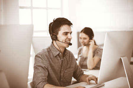 Casual dressed young man using headset and computer while talking with customers online. Group of...