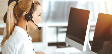 Blond female customer service representative is consulting clients online using headset in sunny...