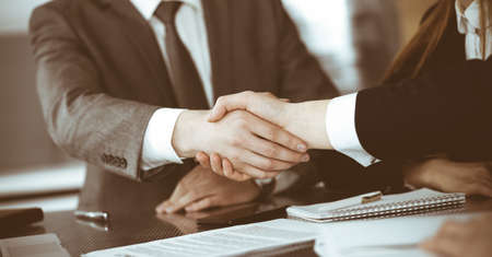 Unknown businessman shaking hands with his colleague or partner above the glass desk in modern office, close-up. Business people group at meetingの写真素材