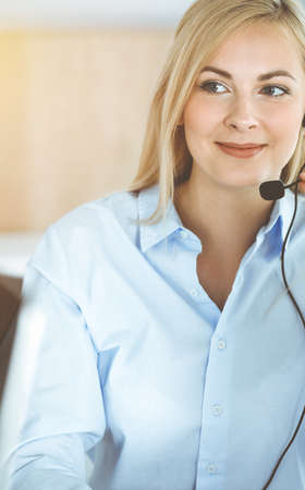 Blonde business woman sitting and communicated by headset in call center in sunny office. Concept of telesales businessの写真素材
