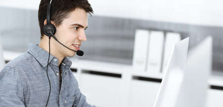 Casual dressed young man using headset and computer while talking with customers online. Call...