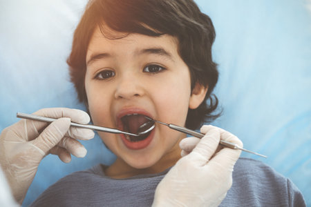 Cute arab boy sitting at dental chair with open mouth during oral checking up with doctor. Visiting...