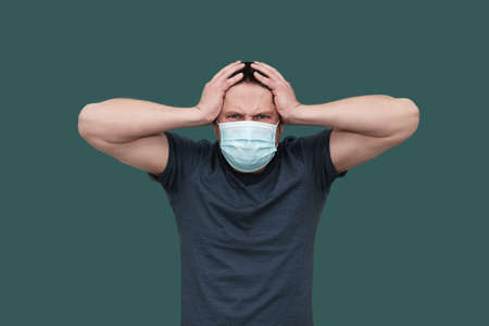 Adult White Man in surgical mask worried, stressed and he holding his head with hands. Isolated on tidewater green color. Care of psychic health. People negative emotions. Tired of Covid-19 pandemic.の写真素材