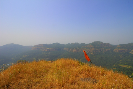View of a flagpole atop a hill with a rugged hilly landscape of the western ghats of Maharashtra, India in the background on a clear day.の写真素材