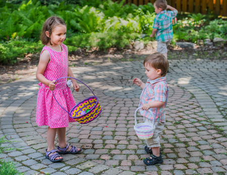 A little boy and girl holding baskets take a break on an Easter egg hunt during the spring season.  A boy in the background holding a basket searches for eggs in a beautiful garden setting. Part of a series.の写真素材