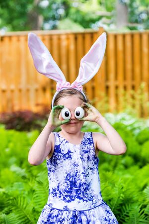 A funny portrait of a girl having fun on Easter wearing bunny ears and holding up silly eyes made from eggs outside in a garden during the spring season.  Part of a series.の写真素材