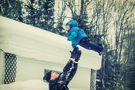 An image captures a happy father throwing his smiling baby son up in the air during a snowfall in the winter season.  Filtered for a retro, vintage look.の写真素材