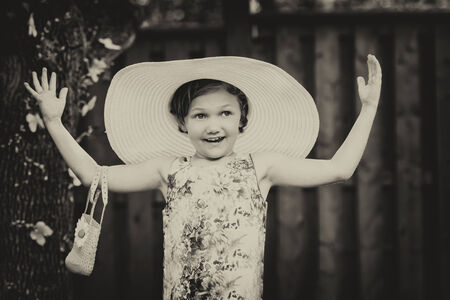 A young girl with a happy expression holds her arms out wearing a white oversized sun hat outside on...