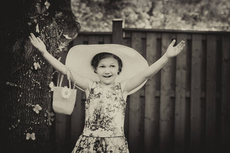 A young girl with a happy expression holds her hands up wearing a white oversized sun hat outside on...