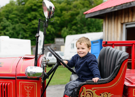 A happy young boy sits in an old shiny vintage red fire truck holding on to the steering wheel...