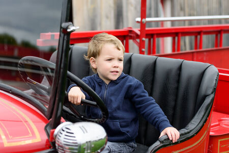 A happy young boy sits in an old shiny vintage red fire truck holding on to the steering wheel...