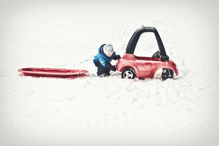 A young boy dressed for cold weather attaches a red sled with a rope to his toy car during the winter season.  Filtered for a retro, vintage look.の写真素材