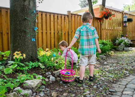 A boy stands holding a basket full of colorful Easter eggs while his little brother searches for...