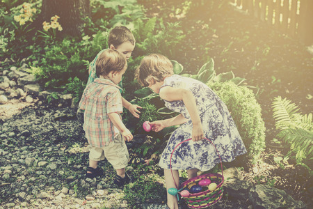 Two boys and a girl search for colorful Easter eggs during an egg hunt outdoors in a garden in the...