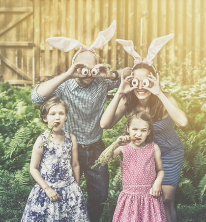 A funny family portrait on Easter of a mother and father wearing bunny ears and holding up silly eyes made from eggs as their children pose eating carrots outside in a garden during the spring season.  Filtered for a retro, vintage look.の写真素材