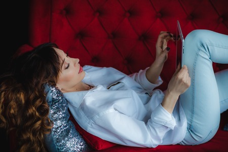 pretty brunette young woman laying on red sofa with tabletの写真素材