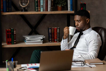 Serious young african businessman drinking coffee while sitting at a table in the office. High...
