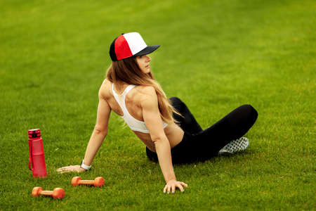 Attractive sporty woman sitting at the grass with dumbbells and water bottle, recommended take daily...