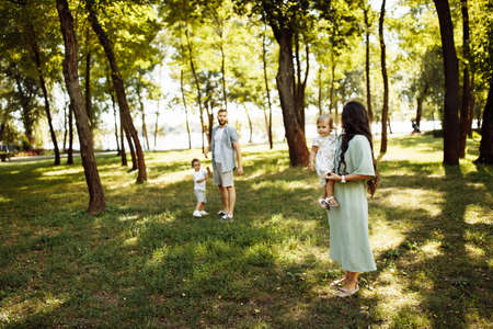 Happy parents with little children at the park, adorable mother hold in arms beautiful daughter, cute son walking with father, happy family weekend, parenting concept. High quality photoの写真素材