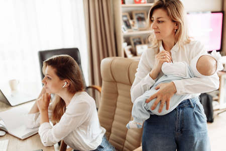 Concentrated woman sit at the desk, looking at the computer, watching webinar training, young mother working at home online, while caring grandmother babysit with newborn baby girl. High quality photoの写真素材