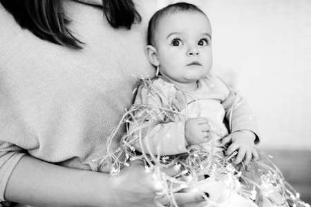 Happy baby girl with light garland sit in mother arms, beautiful toddler celebrate first Christmas, happy family spend winter holidays together at home, xmas concept. High quality photoの写真素材
