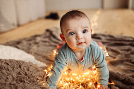 Cute toddler crawl on fours near light garland on blanket, beautiful baby girl excited about first Christmas, new year celebration with little child, winter holidays concept. High quality photoの写真素材