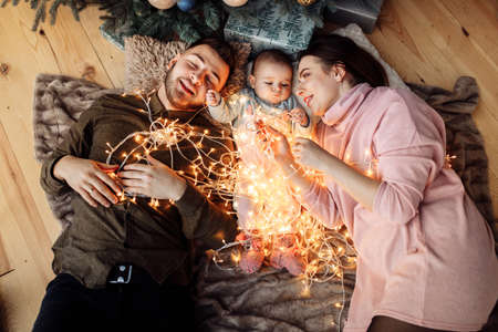 Overjoyed parents lay with baby girl on blanket at home near Christmas tree, happy family celebrate...