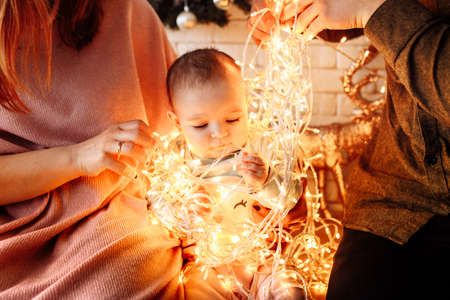Lovely baby girl with light garland sit at the kitchen with caring parents, beautiful toddler...