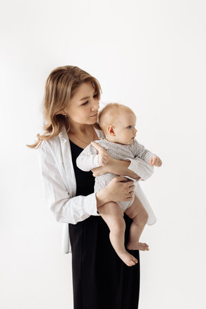 Young mother is holding her lovely daughter in arms and looking to the side with interest. Beautiful attractive woman with cute toddler posing in studio at the white background. High quality photoの写真素材