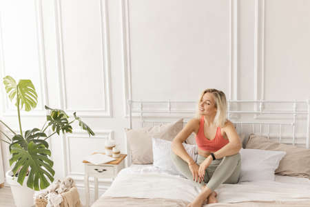 Attractive sportive girl in stylish sportswear sitting on the bed, looking at the side, smiling....