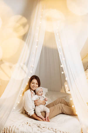 Adorable happy woman with cute baby girl sit on cozy canopy bed, smiling, loving mother hold beautiful little daughter in arms, enjoy tender moments with child, maternity concept. High quality photoの写真素材
