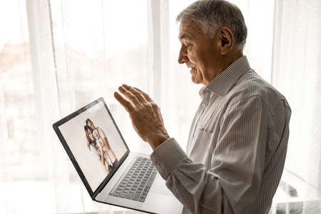 Handsome old man dressed in casual formal style using laptop, while sitting on sofa at home. Happy grandfather communicate with grandchildren. Video call concept. High quality photoの写真素材