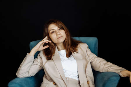 Portrait of stylish beautiful woman sitting on chair in studio. Gorgeous adorable woman in stylish wear posing, looking at the camera. Pretty girl shows female beauty and self confidenceの写真素材