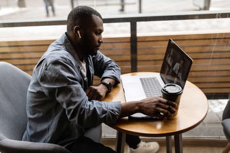 Pensive young employee businessman focused on reading news on a laptop screen, discussing a problem with a colleague in a corporate chat, checking email, holding a glass of tea or coffee in his hand.の写真素材