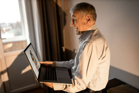 Mature happy man sit on the sofa, looking at the laptop screen. Older male having a video call with grandchildren. Caucasian grandfather use zoom, talking with family by internet, technology conceptの写真素材