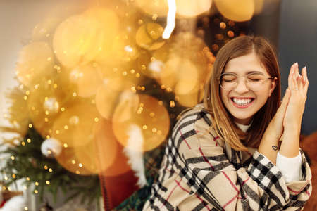 Portrait of smiling lady in glasses sitting near decorated Christmas tree at home, happy woman excited about xmas presents, enjoy New Year celebration, cozy winter holidays concept. High quality photoの写真素材