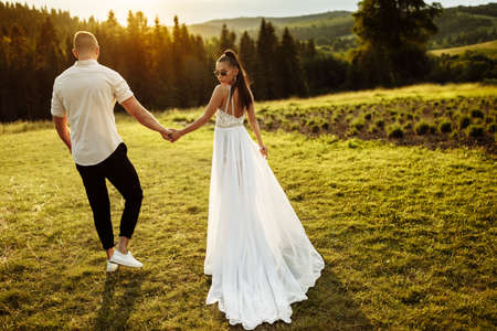 Stylish couple are walking at the park. Handsome groom holding hand of gorgeous bride, enjoy togetherness. Beautiful woman with attractive man on wedding day, posing, photoshoot conceptの写真素材