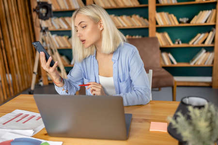 Stylish blonde-hair young lady is using smartphone, browsing internet or chatting with coworkers....