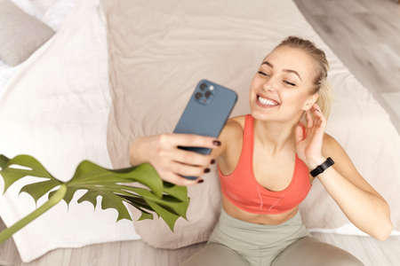 Joyful female fitness trainer is sitting on yoga mat in bedroom, making a selfie or video training....