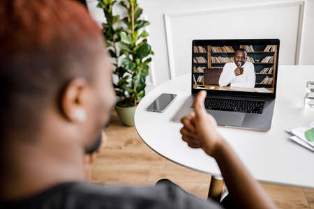 Back view of african american male freelancer showing thumbs up to coworker on video meeting, using...