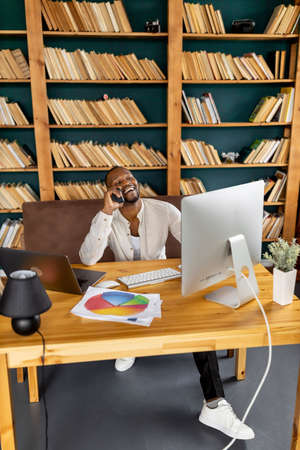 Happy male freelancer sitting at the desk in home office, having a phone conversation with...