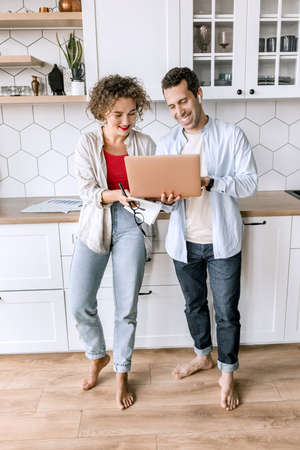 Happy family couple are standing at the kitchen at home, doing online shopping or chatting with friends. Joyful stylish husband and wife using laptop, watching funny videos or photos, smilingの写真素材