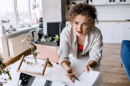 Stylish female freelancer asking an advice from colleague, working on startup, making notes about ideas. Attractive young business woman sitting at the desk in modern office, developing a projectの写真素材