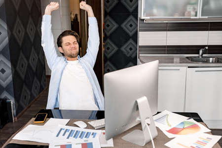 Stylish male employee sitting at the desk in home office, stretching. Caucasian young adult...