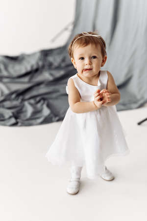 An adorable baby girl thoughtfully looking into camera taking her first steps away from mama, clapping hands, wearing a beautiful dress on the white and gry background. High quality photoの写真素材